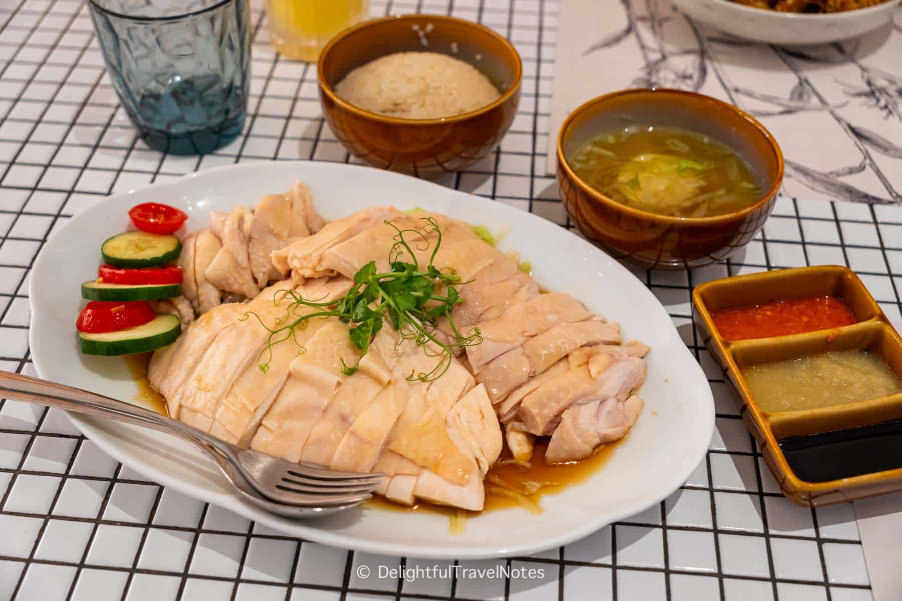 Haianese Chicken Rice at Chatterbox Restaurant in Singapore, with a plate of boneless chicken, bowls of rice, soup and sauces on the table.